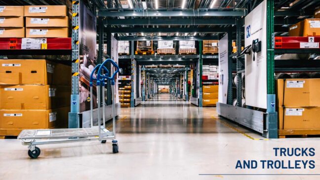 Warehouse aisle with a metal platform trolley in the foreground and stacked boxes on storage racks.